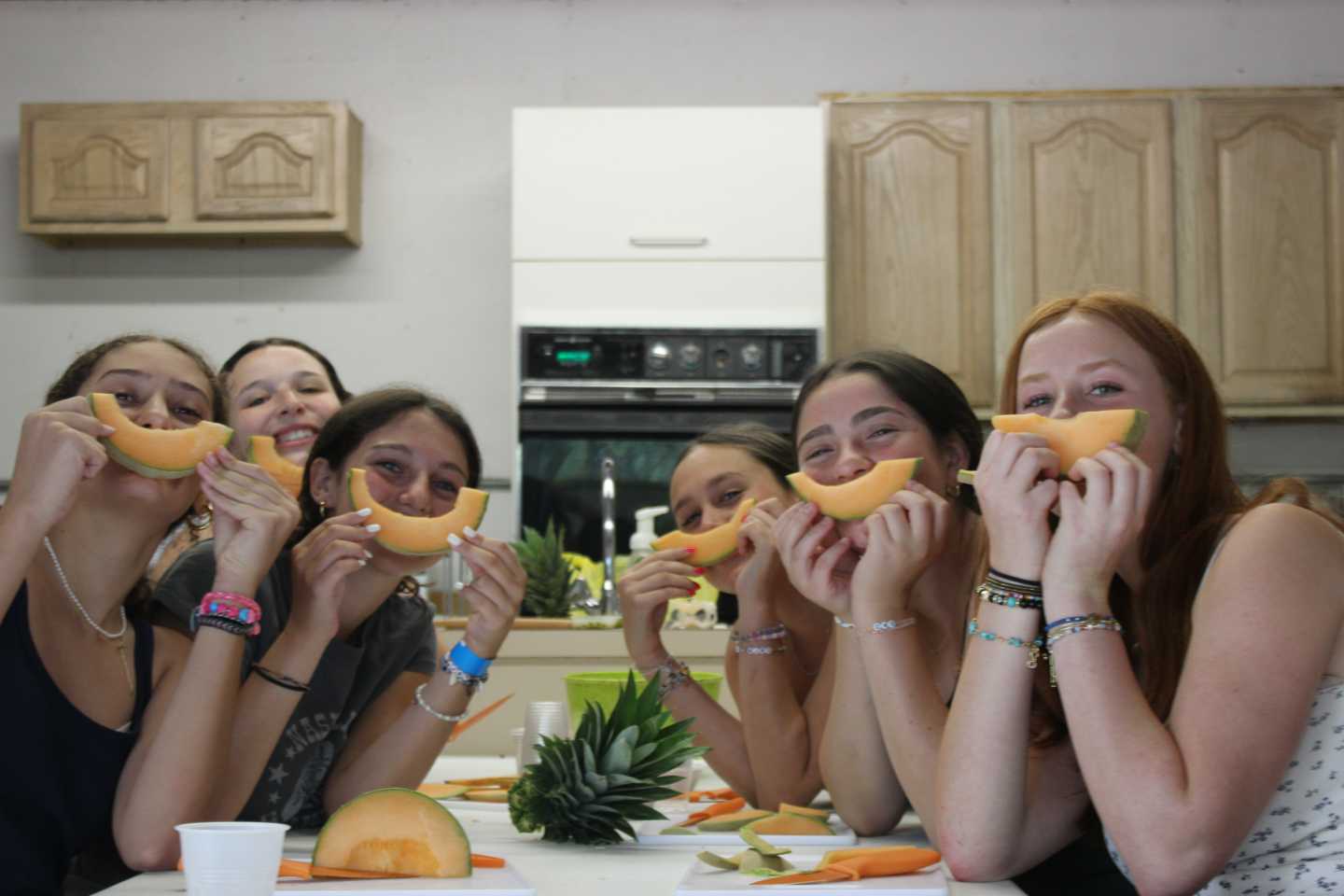 Six people sitting at a table hold slices of cantaloupe in front of their mouths, smiling.