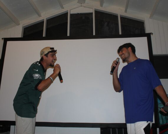 Two men holding microphones face each other and sing in front of a blank projection screen indoors, capturing the lively spirit of a Jewish youth program.