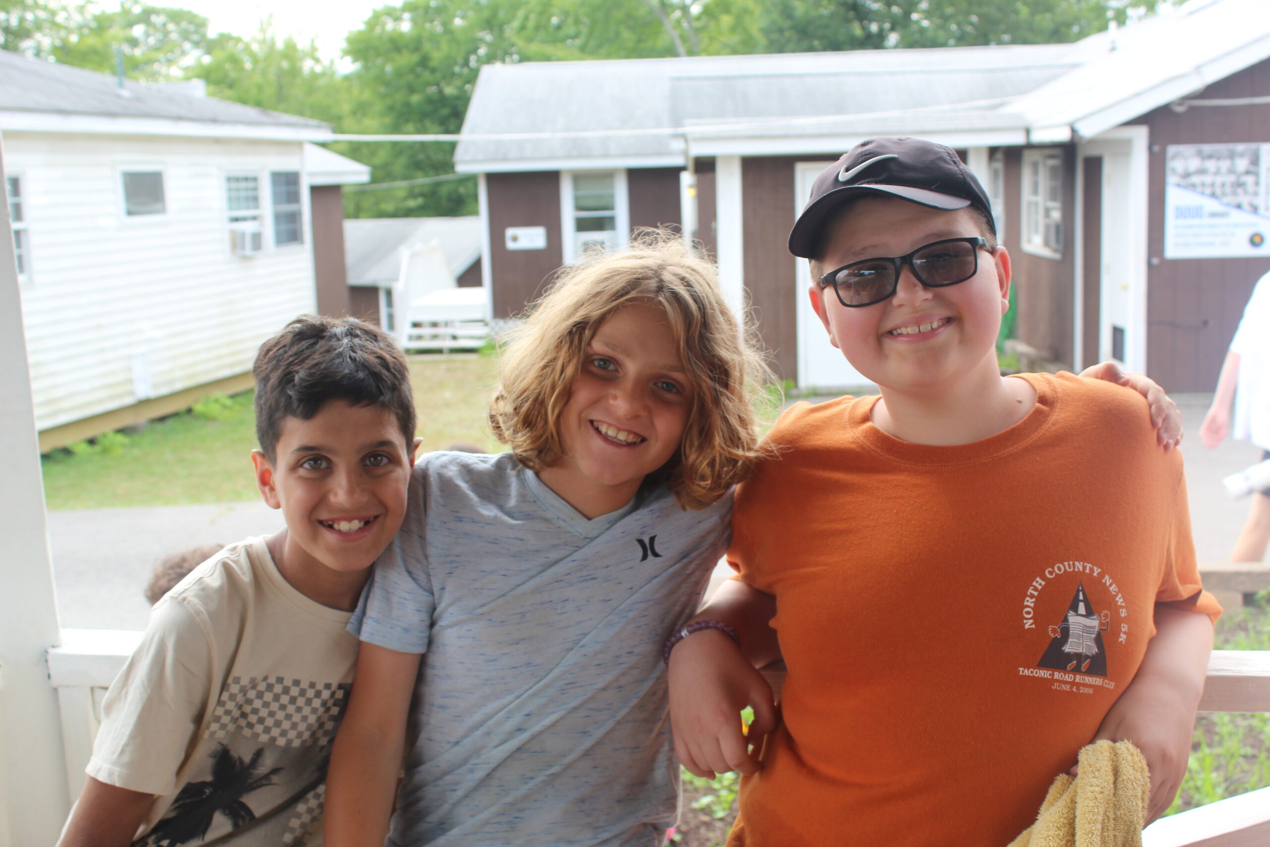 Three smiling children stand close together outside in front of camp cabins, one wearing glasses and a camp shirt, all appearing happy and relaxed at a Jewish summer camp.